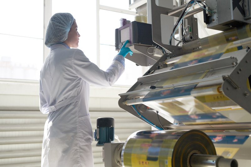 Side view portrait of female factory worker wearing clean white coat standing by machine units at packaging line